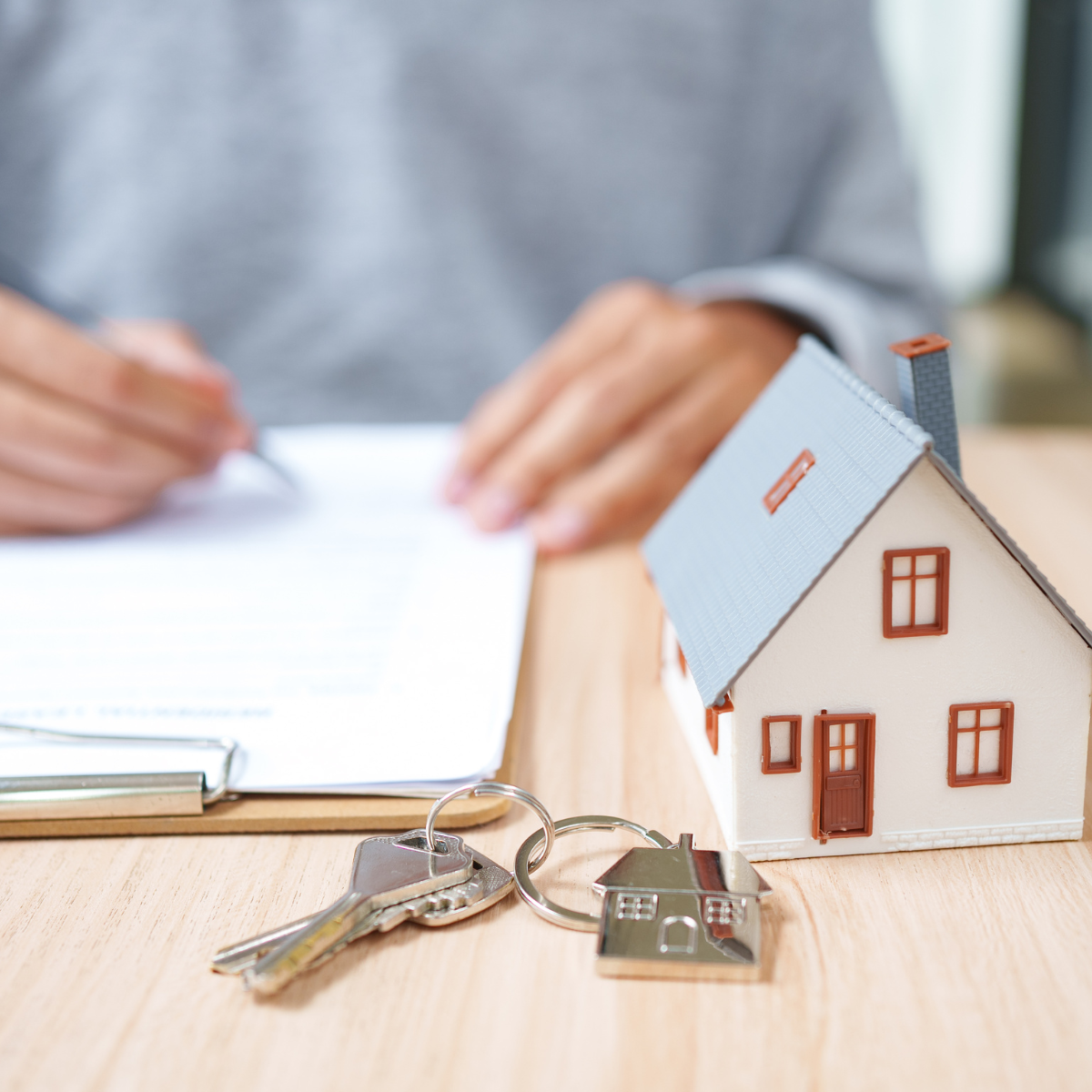 Person signing mortgage documents beside a small model home and house key on a wooden desk.
