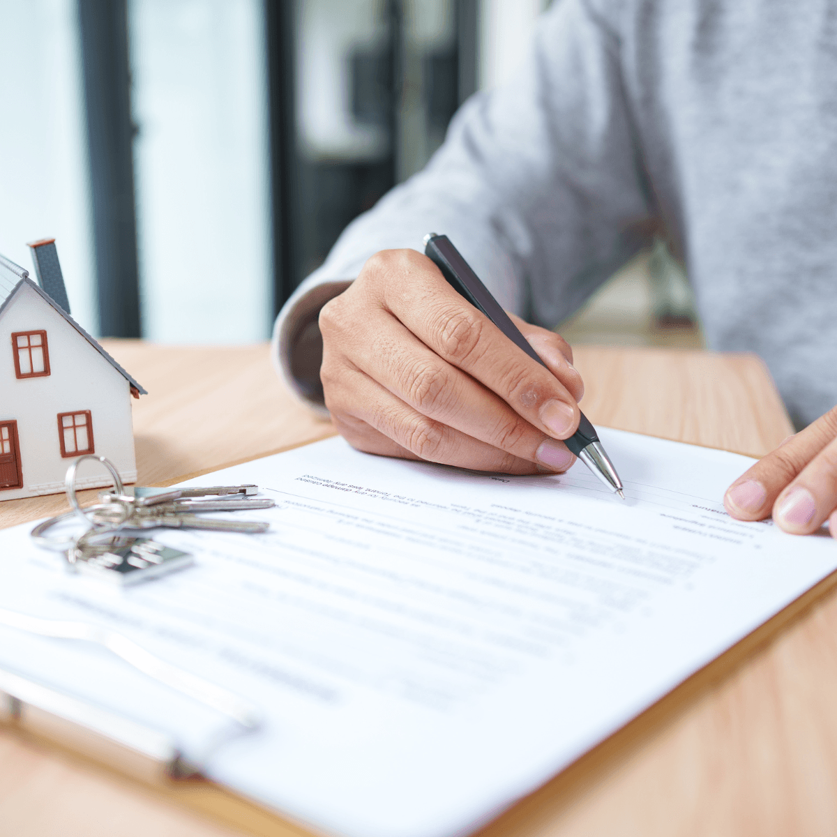 Close-up of a hand signing a loan document with a model home and keys nearby.