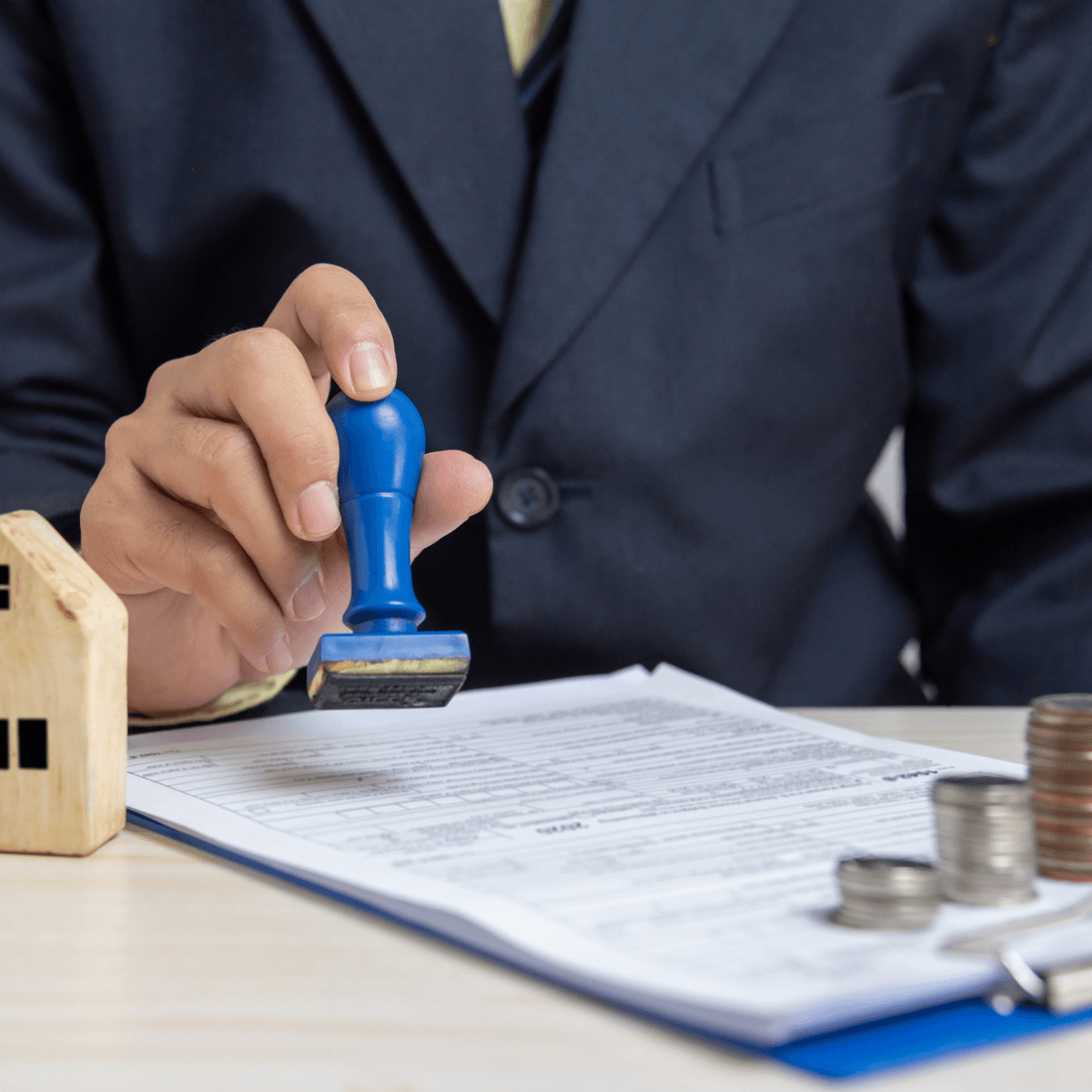 Lender stamping an approved document beside a wooden house model and stacked coins.