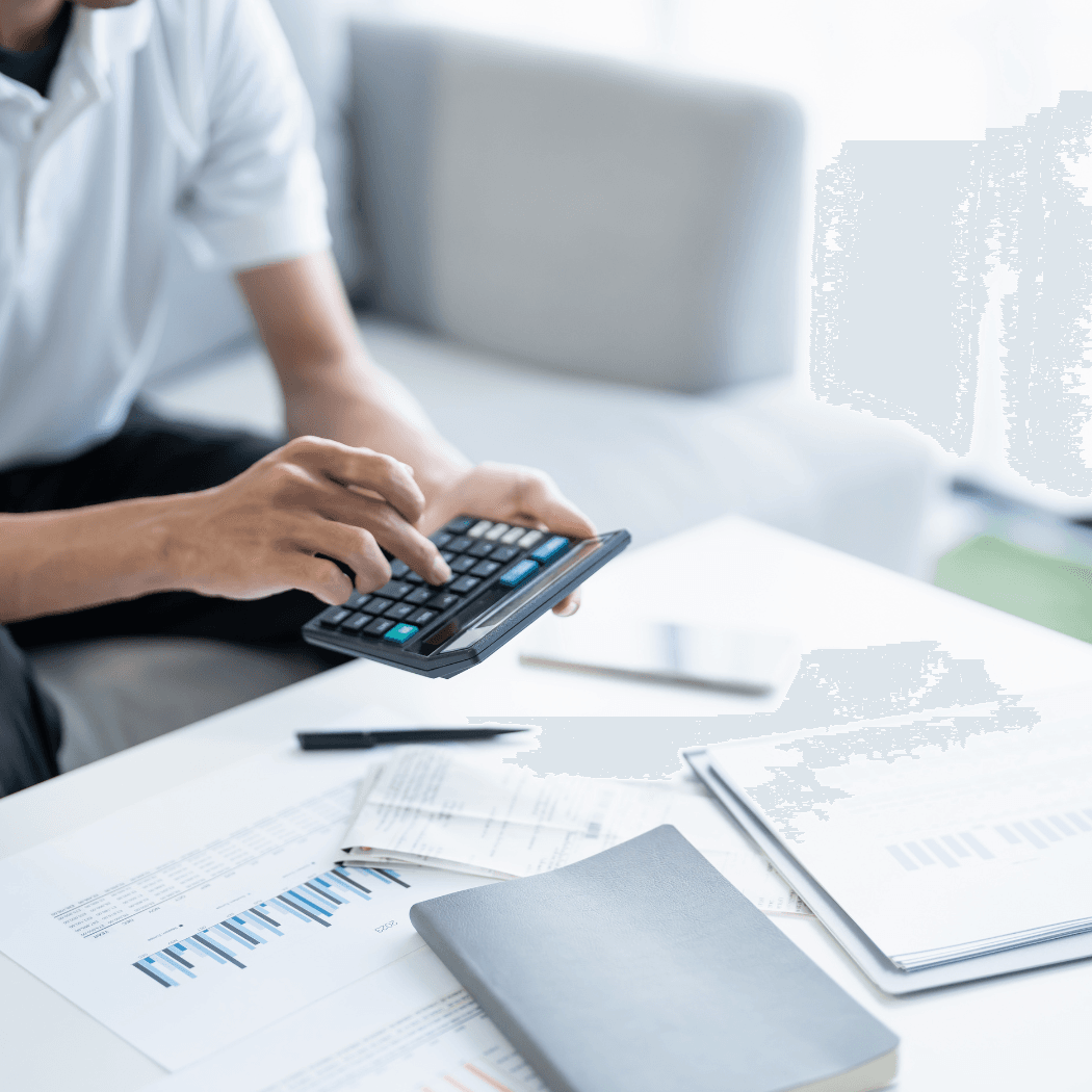 A person in a white shirt using a handheld calculator while reviewing financial charts and documents spread across a white desk, with a laptop in the background.