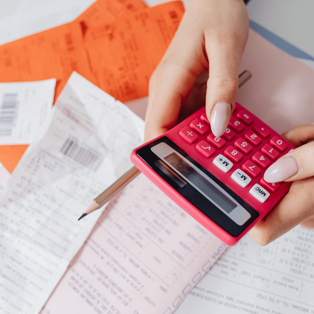 An overhead view of a woman's hand holding a pink calculator and pencil over a pile of receipts, invoices, and financial documents.