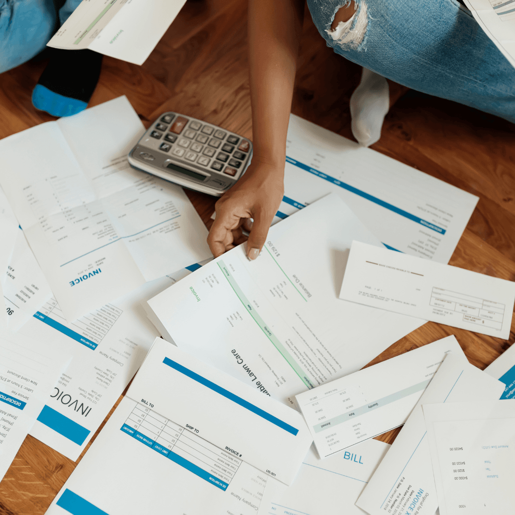 An overhead view of a person sitting on a hardwood floor surrounded by scattered invoices and bills, with one hand resting on a calculator.