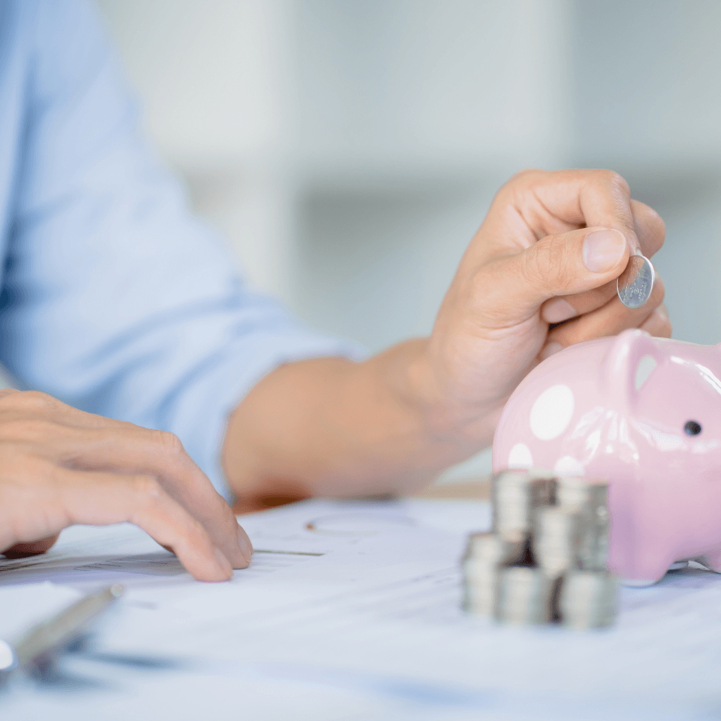 A close-up of a person inserting a coin into a pink ceramic piggy bank, with stacks of coins and financial documents on the desk in front of them.