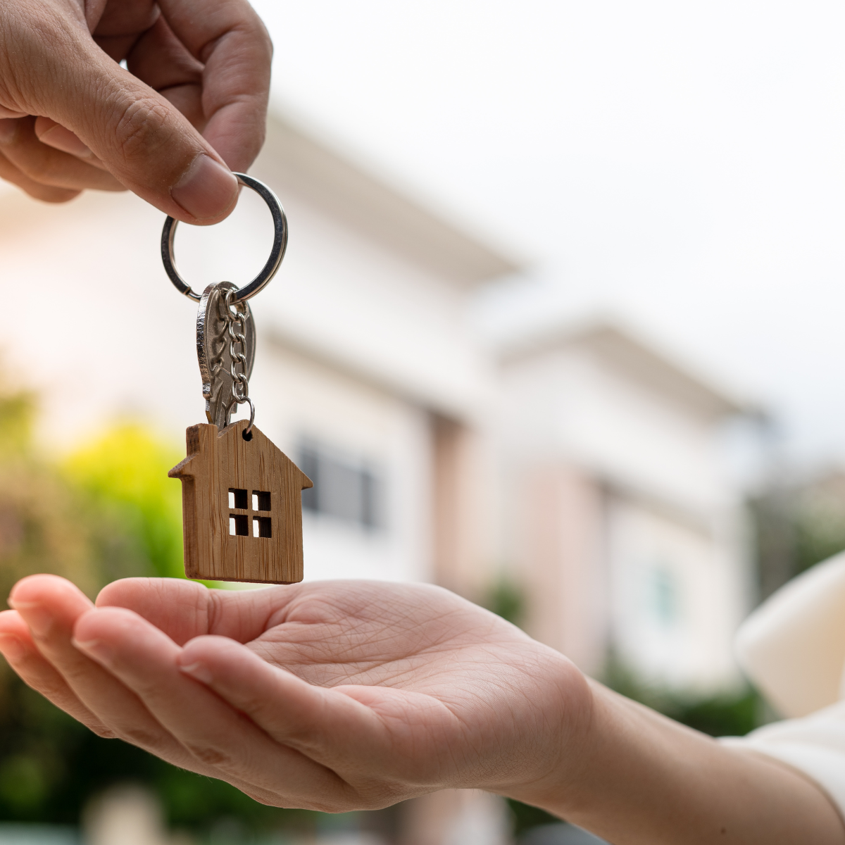 A hand passing a set of house keys with a wooden house-shaped keychain to an open palm, with a blurred residential home in the background.