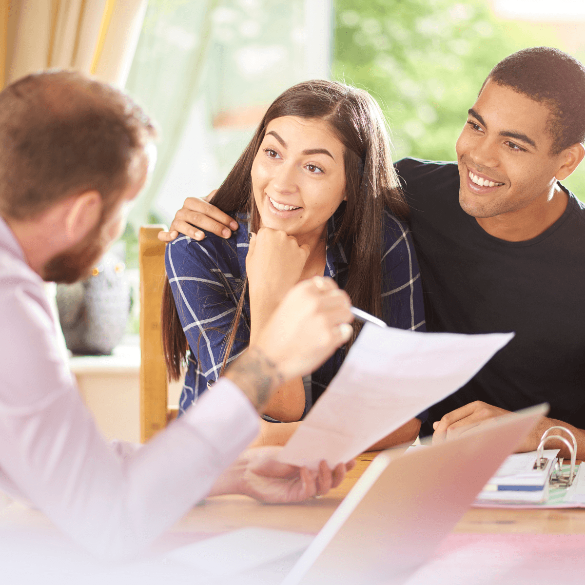 A young couple smiling as they review paperwork with a mortgage advisor across a bright, sunlit table.