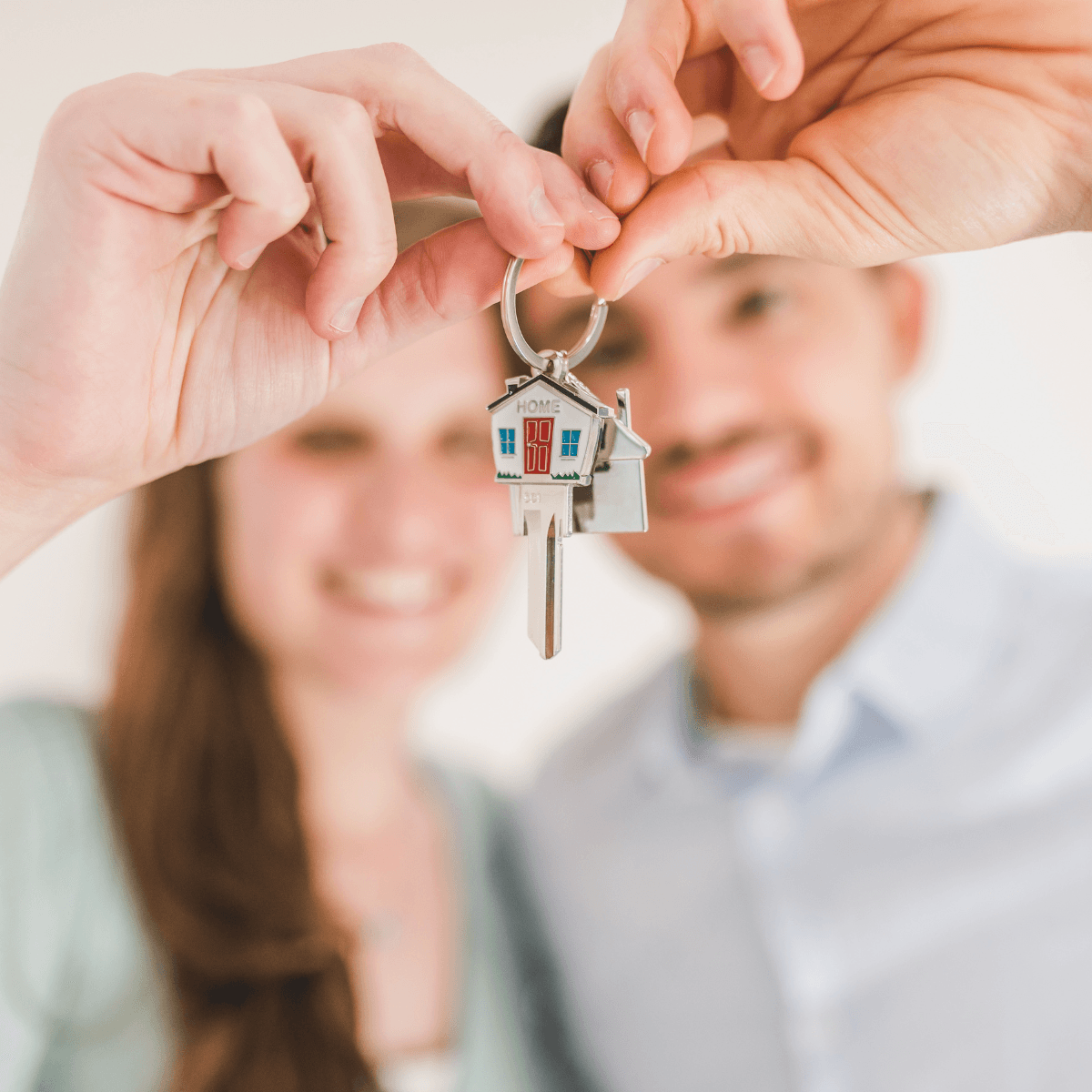A smiling couple holding up a house key with a colourful home-shaped keychain toward the camera, with their faces softly blurred in the background.