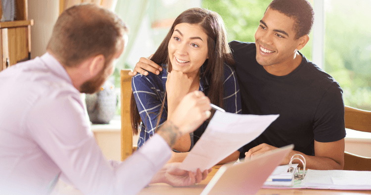 Smiling couple sits close together at a table while an advisor holds a document and points with a pen