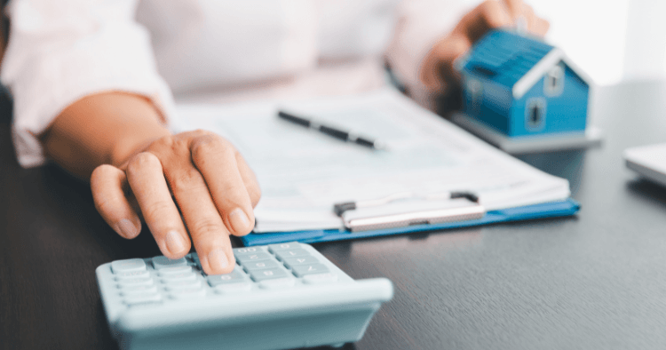 Hand presses calculator keys beside a clipboard of papers and a small blue model house