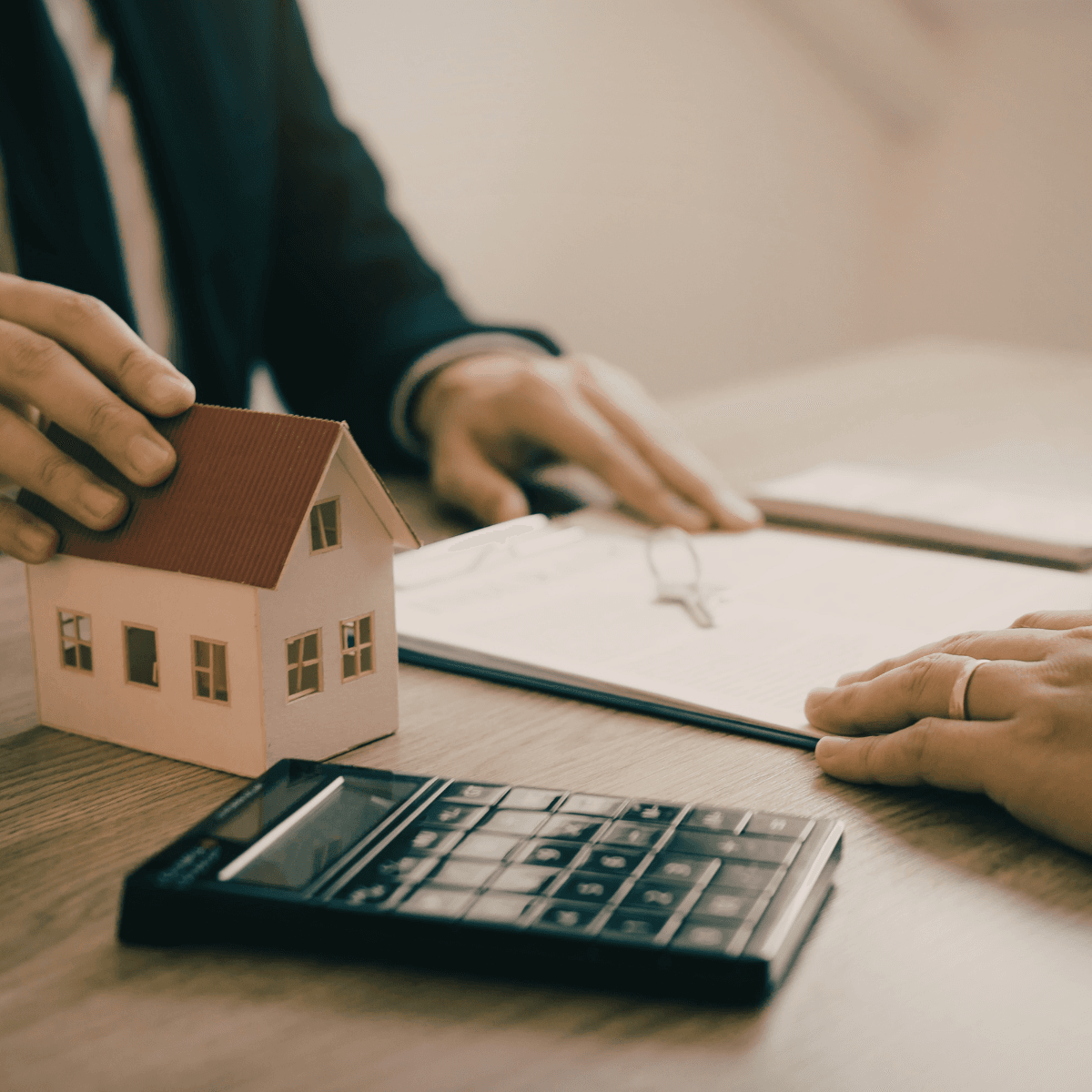 A mortgage advisor holds a small model home on a wooden desk during a client meeting, with a calculator, house keys, and paperwork visible between them.