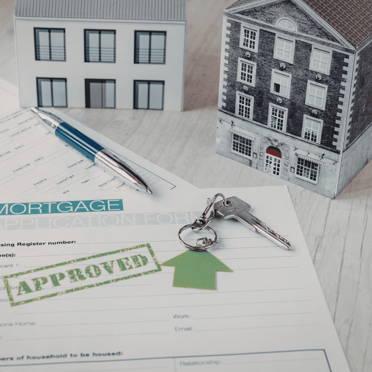 An approved mortgage application form on a desk, with a pen, a set of house keys, and two architectural model homes in the background.