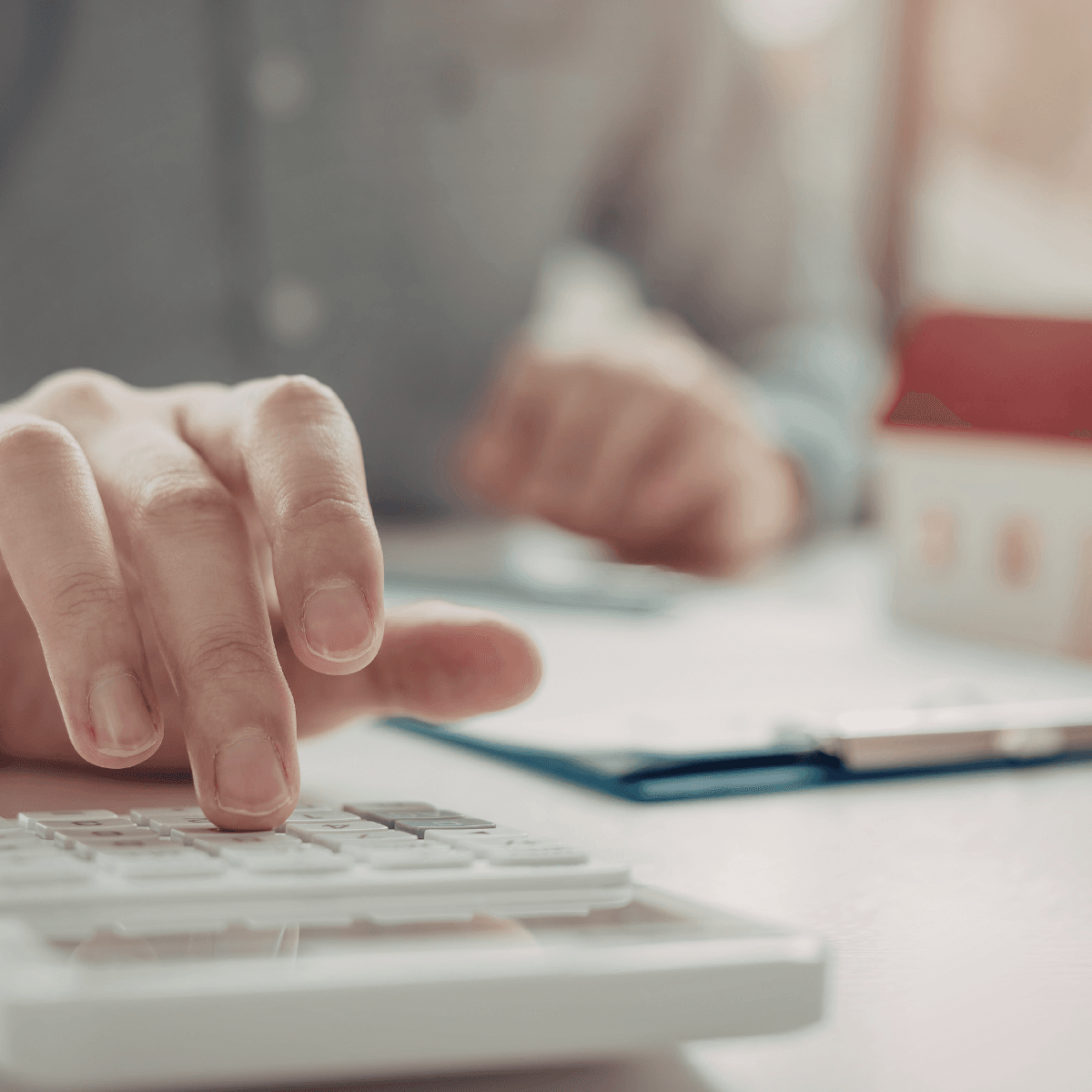 A close-up of a person's hand pressing keys on a calculator at a desk, with mortgage paperwork on a clipboard and a small model home blurred in the background.