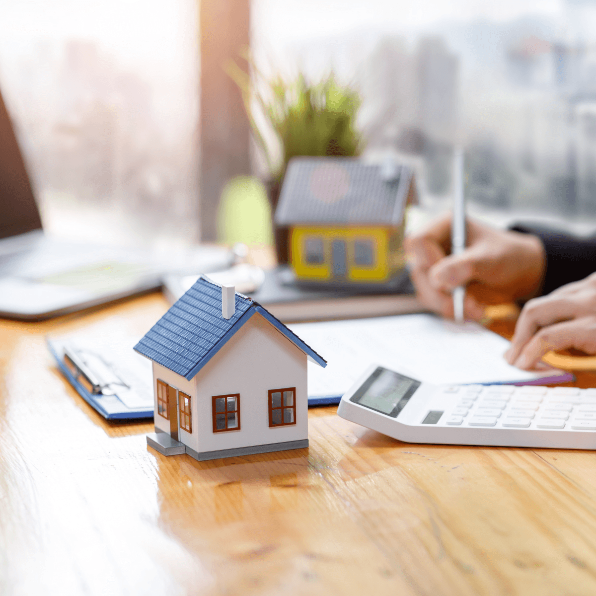 Two small model homes sit on a wooden desk in the foreground while a person writes and works with a calculator in the background.